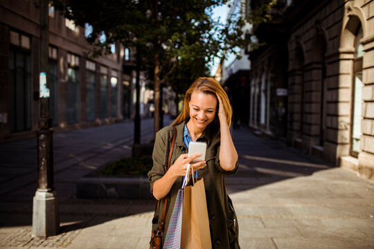 Young adult woman smiling at smartphone with shopping bags downtown
