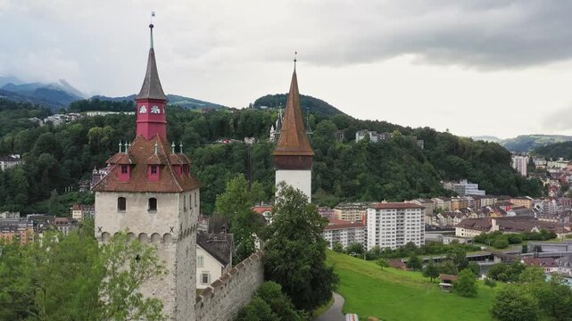 Aerial view of the historic Musegg Wall towers, with their distinctive red and brown roofs, standing tall against the backdrop of lush green hills, lucerne, Switzerland.