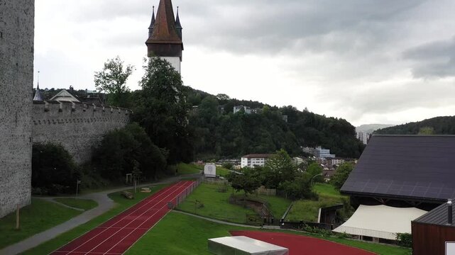 Aerial view of the historic Musegg Wall and its iconic towers amidst a city nestled against lush green hills under a cloudy sky, lucerne, lucerne, Switzerland.