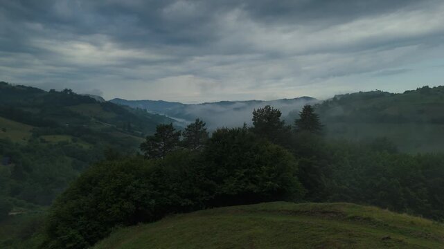 Moody aerial landscape of the Transylvanian hills in Romania, showing a mist filled rural valley, rolling green pastures, hay meadows and scattered farmhouses in the foothills of Carpathian Mountains