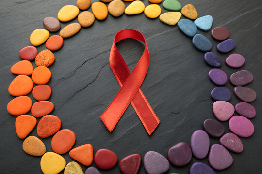 Red ribbon centered in circular rainbow stone arrangement on dark slate background, symbolizing HIV/AIDS awareness and inclusion. generative ai