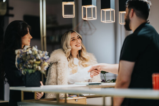 A cheerful blonde woman in a fur coat smiles during a greet at a hair studio reception, shaking hands with a stylist while a friend holds a bouquet in a stylish, modern space.