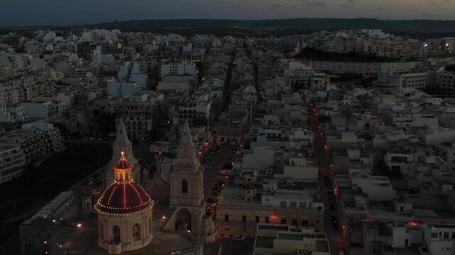 Aerial view of the illuminated Mellieha Parish Church stands majestically amidst the urban sprawl, a beacon of light in the twilight, Mellieha, Malta.