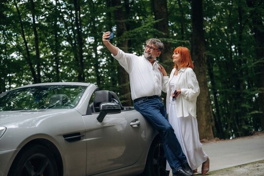 Doing the selfie. Lovely senior couple standing on the road near convertible cabriolet car