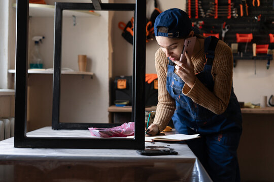 craftsman repairwoman is making notes speaking on mobile phone in workshop on tools background. The concept of industrial and work environment.