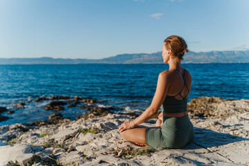 Woman meditating in nature with sea view. Young female practicing yoga in beach. Beautiful woman exercising outdoors and practicing yoga asanas at seaside. Holistic Health and Mental Well-Being.