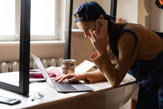 Female carpenter in blue overalls working in her workshop talking on smartphone taking order and using laptop computer at workplace