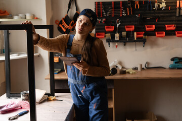 Young female carpenter in blue uniform holding tablet standing near metal table legs in workshop