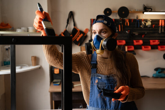 handyman woman worker wearing protective workwear painting with black paint metal table legs at workshop