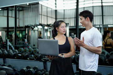Asian man and woman exercise training in fitness gym. Young female trainer using laptop for schedule exercise training to fitness male. Workout training in fitness gym.