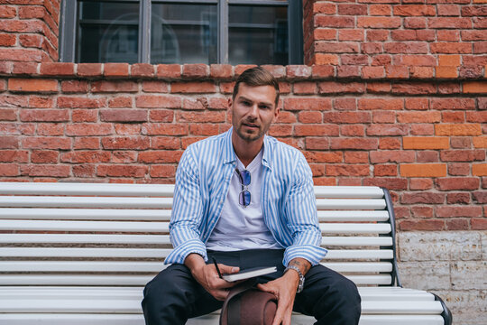 A contemplative man in a striped shirt sits on a white bench against a red brick wall, holding a book and hat, exuding a relaxed and thoughtful vibe. - Powered by Adobe