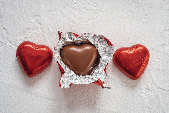 Heart-shaped chocolates wrapped in shiny red foil are arranged on a light-colored surface. One chocolate is open, showing a tasty chocolate heart inside.