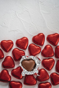 Heart-shaped chocolates wrapped in shiny red foil are arranged on a light-colored surface. One chocolate is open, showing a tasty chocolate heart inside.