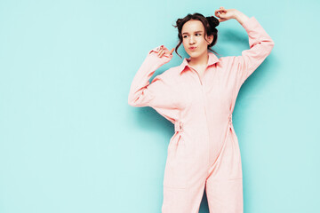 Young beautiful smiling female in trendy summer pink overalls. Sexy carefree woman posing near blue wall in studio with two horns. Positive model having fun and going crazy. Dancing