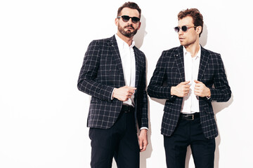 Portrait of two handsome confident stylish hipster lambersexual models. Sexy modern men dressed in black same elegant suit. Fashion male posing in studio near white wall. In sunglasses