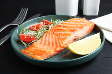 Piece of tasty grilled salmon, lemon, tomato slices and microgreens served on black wooden table, closeup