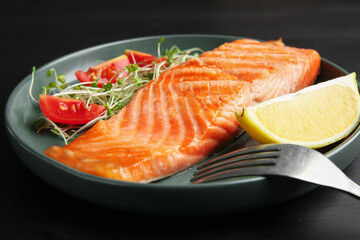 Piece of tasty grilled salmon, lemon, tomato slices and microgreens served on black wooden table, closeup