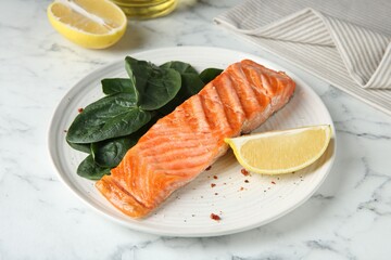 Piece of tasty grilled salmon, lemon slice and spinach served on white marble table, closeup