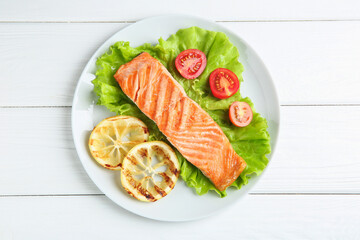 Piece of tasty grilled salmon, lemon slices and salad on white wooden table, top view