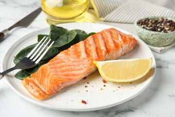 Piece of tasty grilled salmon, lemon slice and spinach served on white marble table, closeup