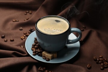 Aromatic coffee in cup, beans and brown sugar on fabric, closeup
