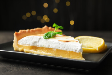 Slice of tasty lemon meringue tart with mint served on black table against blurred lights, closeup