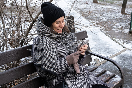 Woman in warm winter coat and hat sitting on a park bench, smiling while actively using a modern smartphone, illustrating digital connectivity and social media use in the cold season