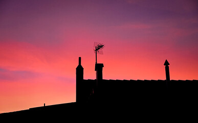 Silhouette of a rooftop with chimneys and a TV antenna against a vivid red and purple sunset sky, creating a dramatic and peaceful evening atmosphere