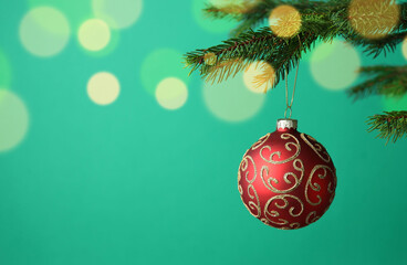 Red Christmas ball with pattern hanging on fir tree branch against green background with blurred lights, closeup with space for text. Bokeh effect