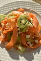Fresh carrot ribbon salad with kiwi and mandarins sprinkled with dried herbs and cheese crumbs on a white plate, top view