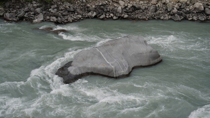 Large gray boulder partially submerged in rushing turquoise-white water and foam, geology concept.