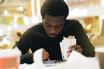 Young african male eating doner fast food in cafeteria while using smartphone