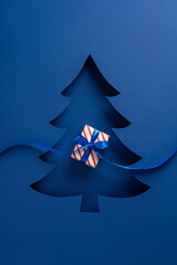 A vertical blue greeting card features a layered Christmas tree cutout with a small striped gift...