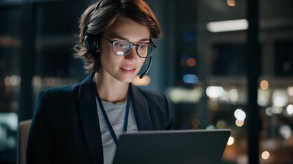 Focused businesswoman wearing headset working on laptop in modern urban office at night - Powered by Adobe