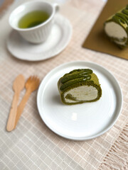 A slice of matcha roll cake served on a white ceramic plate with green tea and wooden cutlery on a beige cloth.Bright,clean tabletop food photography ideal for dessert menus and bakery promotions.