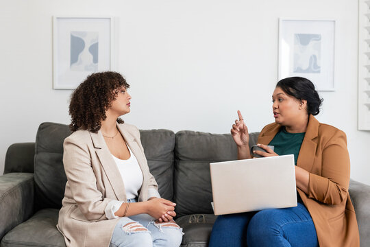 Diverse female colleagues talking in living room on gray sofa using silver laptop with coffee mug