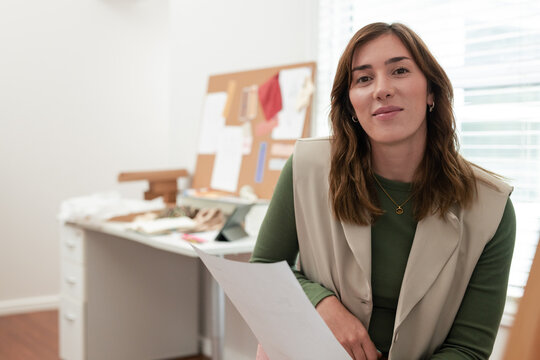 Female designer holding paper and examining corkboard sketches at desk in home studio, copy space