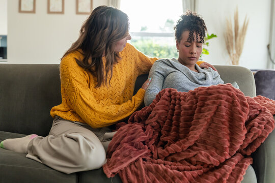 Diverse female friends sitting on living room sofa, one wrapping in rust blanket by window, art