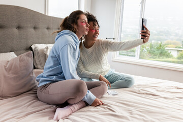 Female friends sitting on cozy bed wearing under-eye patches and taking selfie with smartphone