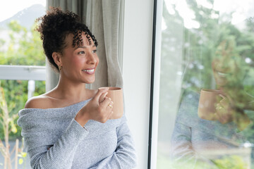 Woman in her 30s standing by window at home holding ceramic mug wearing off shoulder sweater