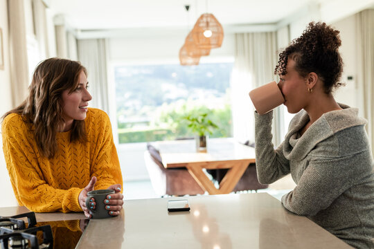 Diverse female friends holding coffee mugs and chatting at kitchen island near smartphone