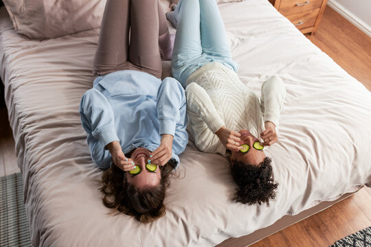 Diverse female friends lying on large bed wearing loungewear and holding cucumber slices over eyes