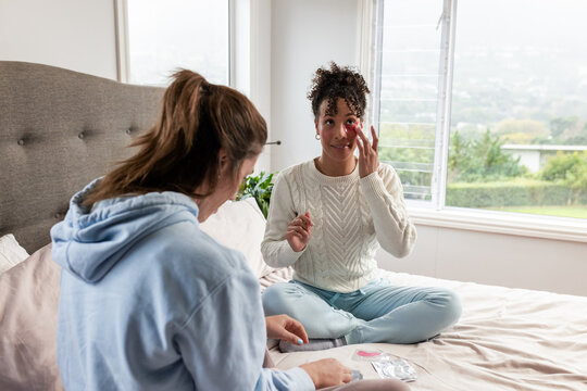 Diverse female friends applying under-eye patch, sorting cosmetics on bed at home, copy space - Powered by Adobe