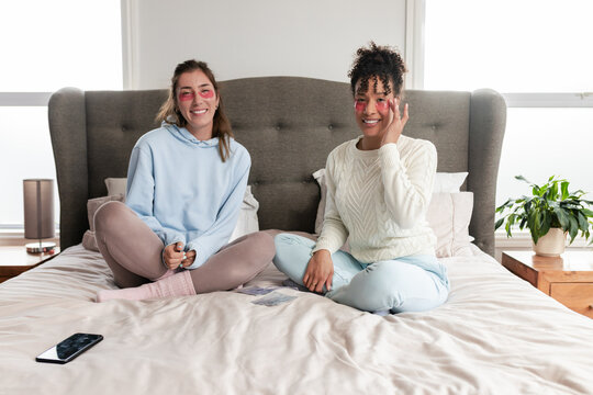 Diverse female friends applying under-eye gel patches while sitting cross-legged on king-size bed - Powered by Adobe