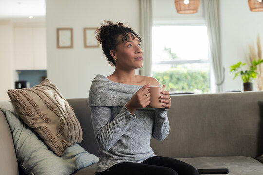 African American woman sitting on gray sofa in living room holding beige mug gazing out window