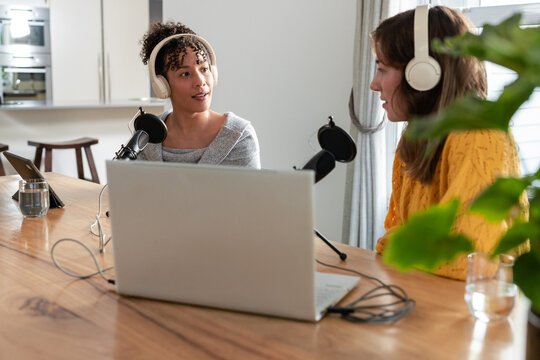 Diverse female coworkers recording podcast in kitchen studio using microphones and laptop