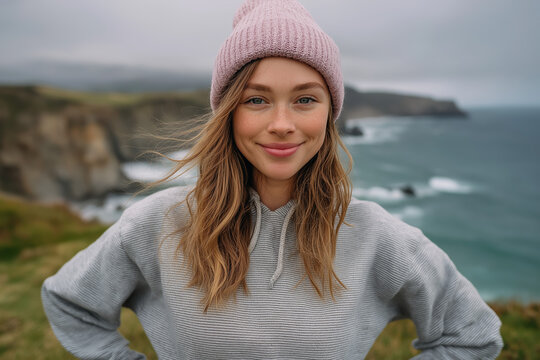 Cheerful portrait of woman in pink knitted beanie standing near ocean cliffs on cool overcast coastal day

