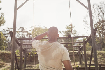 Man standing centered at park, facing wooden obstacle structure with ropes and damp sweat on shirt