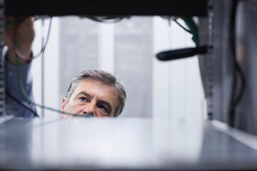 Mature adult male technician leaning into server rack, inspecting bundled cables and components