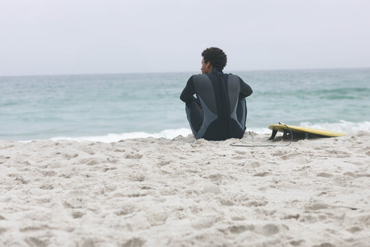 African American man sitting on beach in dark wetsuit facing ocean near yellow surfboard leash - Powered by Adobe
